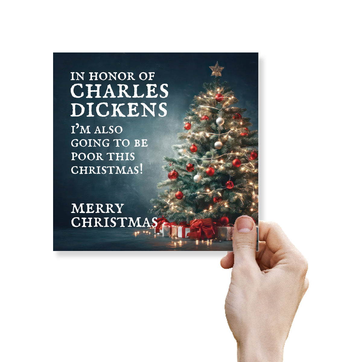 A hand holds a square Christmas greeting card against a plain white background. The card features a dark blue background with white text reading 'IN HONOR OF CHARLES DICKENS I'M ALSO GOING TO BE POOR THIS CHRISTMAS! MERRY CHRISTMAS!' and a decorated Christmas tree with red ornaments and wrapped gifts.