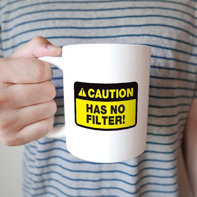 Person holding a white mug with a yellow and black caution label reading 'CAUTION HAS NO FILTER!' against a striped background.