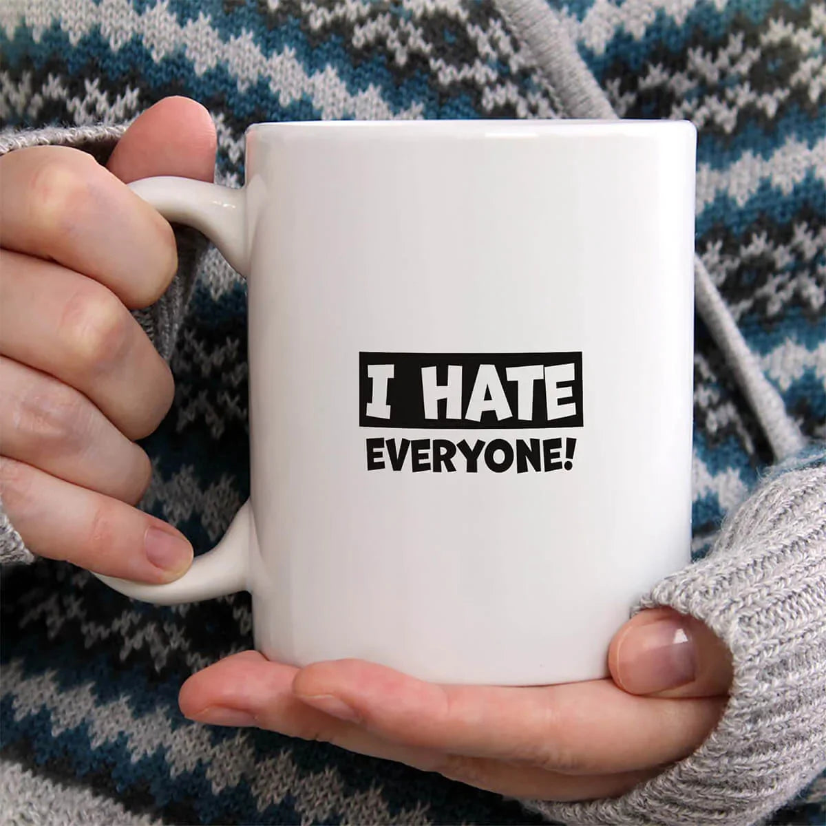 White mug with 'I HATE EVERYONE!' text held by a person wearing a patterned sweater.
