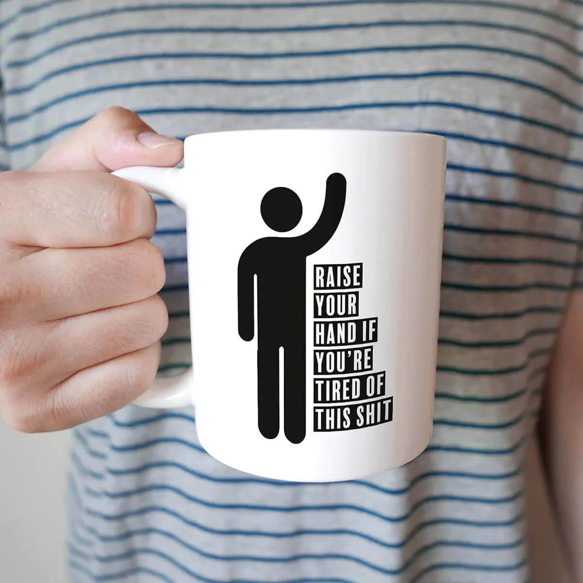 Person holding a white mug with a black silhouette and text design, set against a striped shirt background.