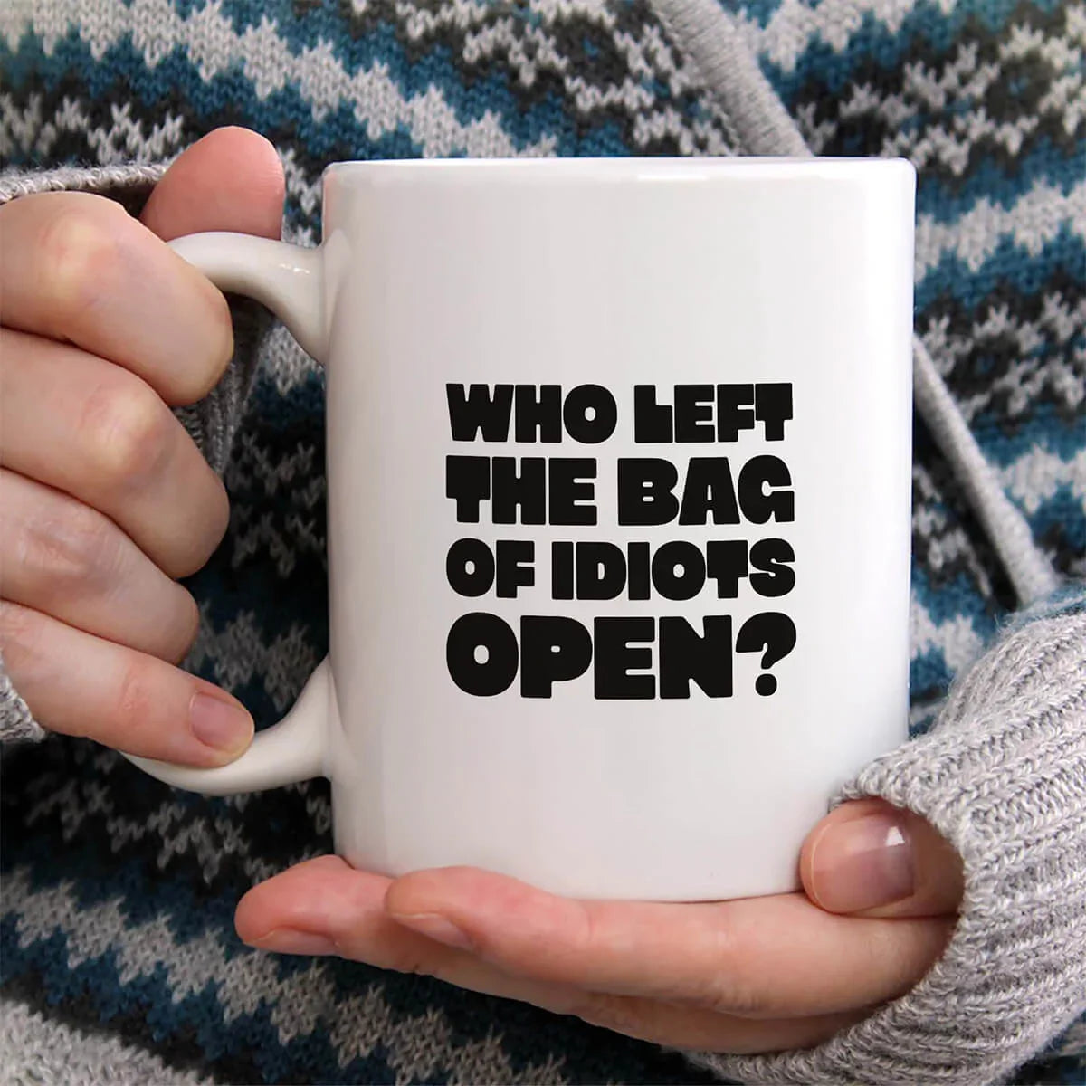 Person holding a white mug with text 'WHO LEFT THE BAG OF IDIOTS OPEN?' against a patterned fabric background.