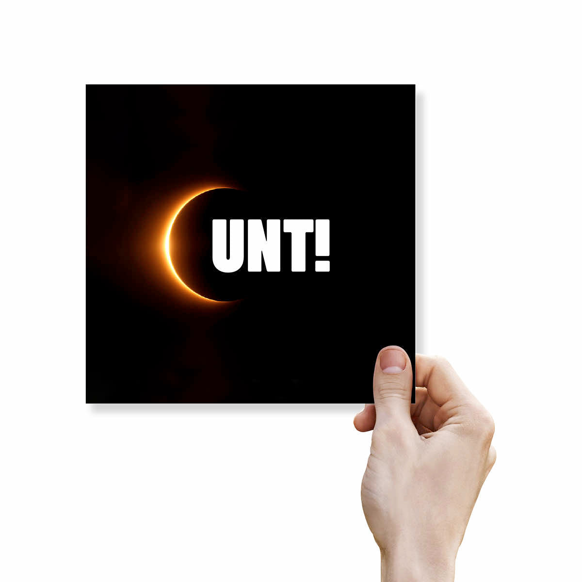 A hand holding a square black art print or coaster featuring a glowing orange eclipse and the bold white text 'UNT!'.